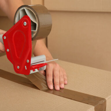 Eine Person verschließt mit einem roten Handabroller einen Umzugskarton mit braunem Klebeband