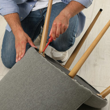 Eine Person befestigt mit einem Schraubendreher die Holzbeine an einem grauen gepolsterten Stuhl.