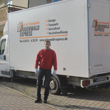 Ein Umzugsmitarbeiter steht vor einem weißen Transporter mit der Aufschrift ‚Odenwald Express – Grab Transporte‘, der am Straßenrand geparkt ist.