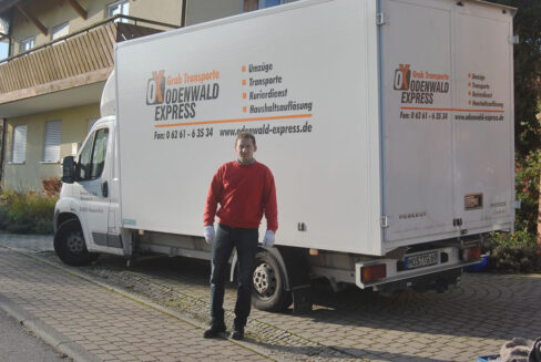 Ein Umzugsmitarbeiter steht vor einem weißen Transporter mit der Aufschrift ‚Odenwald Express – Grab Transporte‘, der am Straßenrand geparkt ist.
