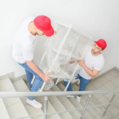 Zwei Umzugshelfer tragen ein mit Folie verpacktes Möbelstück vorsichtig ein Treppenhaus hinunter.
