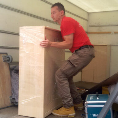 Eine Person mit weißen Handschuhen verpackt ein großes Möbelstück sorgfältig mit transparenter Schutzfolie, um es für den Transport zu sichern.